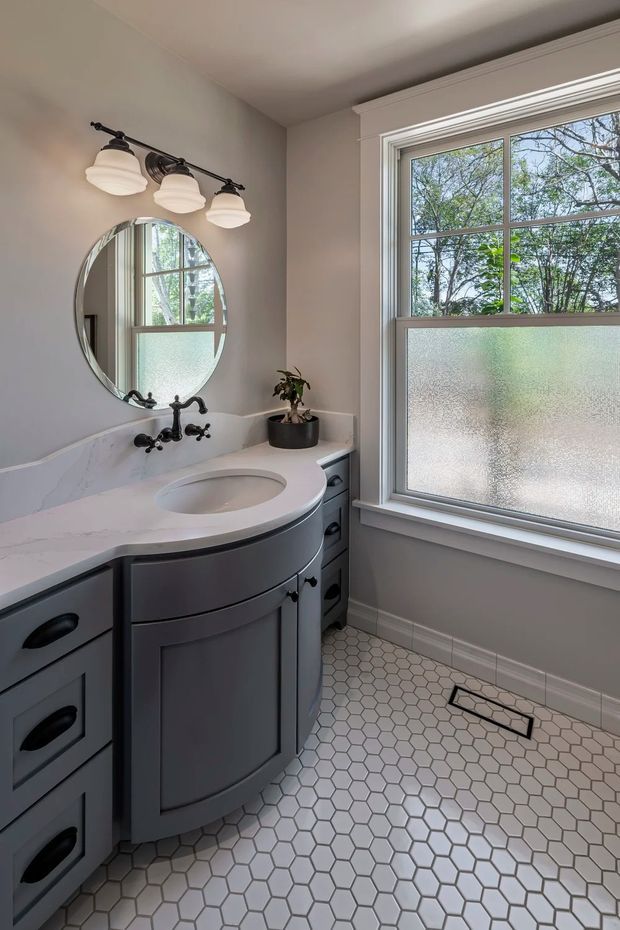 A compact, traditionally styled bathroom featuring a curved dark grey vanity with integrated white countertop and undermount basin, black vintage-style faucet hardware, marble-look backsplash, round wall mirror, and three-light wall sconce. The room has a large frosted-glass window with white trim and a white hexagonal tile floor with dark grout.