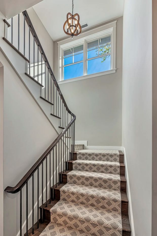 A bright interior stairway with a winding staircase, dark stained timber treads, slim black metal balusters, and a dark wood handrail. The stairs are covered by a beige and taupe geometric patterned runner. White walls and trim surround a tall window with traditional divided panes, while a sculptural bronze pendant light hangs overhead.