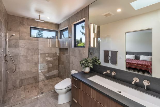 Contemporary bathroom with a large tiled walk-in shower, clear glass screen, wall-hung shower fittings, toilet, and a long timber vanity with a dark stone benchtop and integrated white basin. A large mirror reflects an adjacent bedroom, while skylight and windows provide abundant natural light. The bathroom features warm grey stone-look tiles, dark timber cabinetry, black fixtures and refined modern detailing.