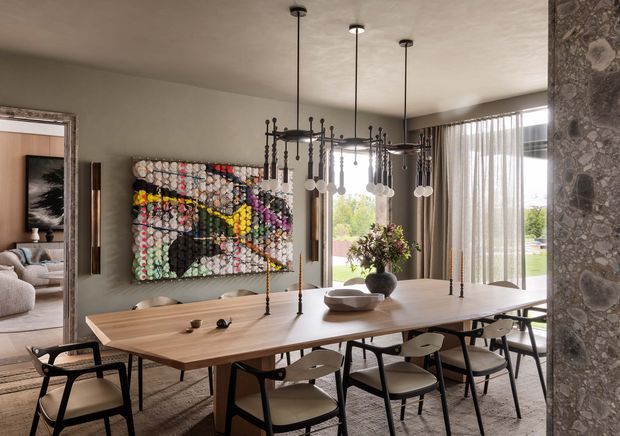 A refined dining room centered on a long light-wood dining table with upholstered and dark-framed dining chairs. Multiple sculptural pendant lights hang above the table, while a colourful abstract artwork provides a strong focal point on the plaster wall. Floor-to-ceiling sheer curtains open to a bright garden view, and a textured stone-clad structural wall frames the right side. A living room is visible through the opening on the left.