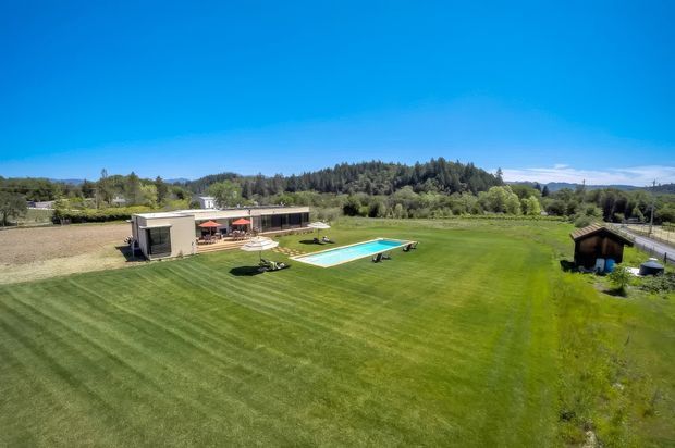 Aerial view of a contemporary single-storey property set within expansive, meticulously mown lawns. The low-profile building has a long horizontal form, with an outdoor entertaining area and a rectangular swimming pool in front, surrounded by open rural landscaping and tree-covered hills.