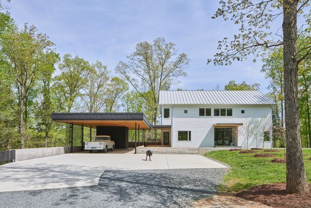 Exterior view of a contemporary two-storey white-clad house set among mature trees. The building has a simple gable roof with standing-seam metal roofing, horizontal wall cladding, dark-framed windows, and a covered glazed entry or patio. A separate flat-roofed carport with dark screening and a timber-toned ceiling sits beside a broad concrete driveway.