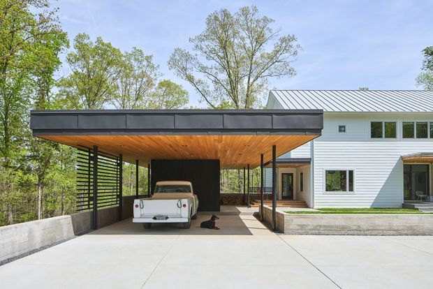 A contemporary residential exterior with a broad flat-roofed carport attached to a white horizontal-clad house. The carport has a warm timber soffit, dark metal fascia, slender dark posts, and horizontal privacy screening, with a vintage car parked beneath. Concrete paving, raised planters, and mature trees surround the property.