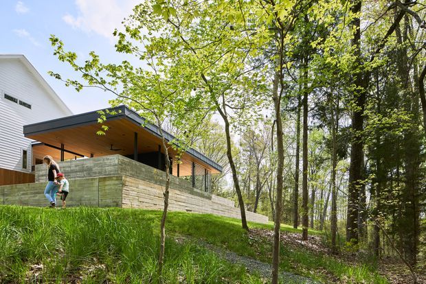 Exterior view of a contemporary, low-profile house addition or pavilion set into a wooded, sloping site. The building has dark metal roof edges, warm timber soffits, extensive open glazing, and layered horizontal concrete or stone retaining walls. Mature deciduous woodland surrounds the structure, with a grassy bank in the foreground and people providing scale.
