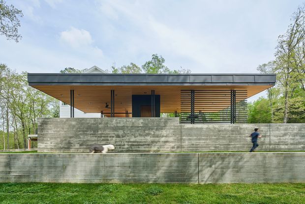 Front exterior view of a low, single-storey contemporary house with a broad flat roof, deep timber-lined eaves, dark framed glazing, horizontal timber screening, and a substantial raw concrete retaining wall. The building is set among mature trees and landscaped grass, with a pedestrian providing scale.