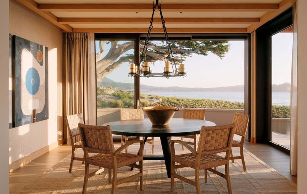 A warm, light-filled dining room with a round dark dining table and woven timber-framed chairs beneath a sculptural chandelier. Exposed timber ceiling beams, timber flooring, full-height glazing, and a large dark-framed glazed door open the room to a tranquil waterfront outlook and landscaped exterior. Abstract wall art and neutral curtains add understated decor.