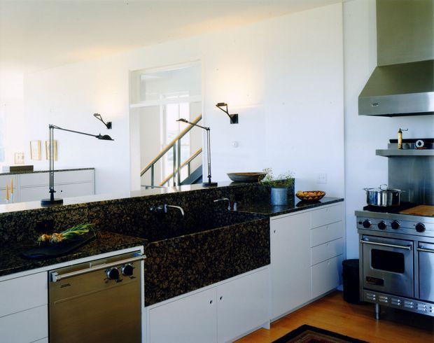 Bright contemporary kitchen with extensive dark speckled stone benchtops and matching splashback, white flat-panel cabinetry, stainless-steel appliances, and timber flooring. A stairway is visible through the central opening, with black articulated wall lighting and a raised breakfast or preparation counter in the foreground.