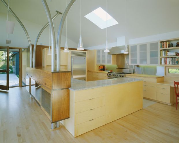 Bright open-plan kitchen with extensive pale timber cabinetry, a large central island, stainless-steel appliances, and a light stone benchtop. Curved exposed metal supports rise through the island and cabinetry, while pendant lights and a rectangular skylight provide illumination. Glazed doors open to an outdoor area, and open shelving displays books and objects at the right.