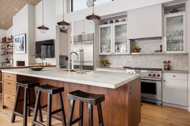 Bright, open-plan kitchen with a large white stone island, warm timber cabinetry and flooring, white shaker-style wall cabinets, glass-front display cupboards, stainless-steel range cooker, tiled splashback, and three pendant lights. The island includes seating for three or more stools, with open shelving and a television visible in the adjacent living area.