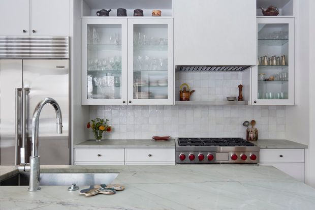 Bright, refined kitchen featuring white shaker-style cabinetry, glass-front display cupboards, a stainless-steel refrigerator and professional range, pale tiled backsplash, and a large pale stone island with an undermount sink and gooseneck faucet. The room has abundant built-in storage and a clean, softly traditional design.