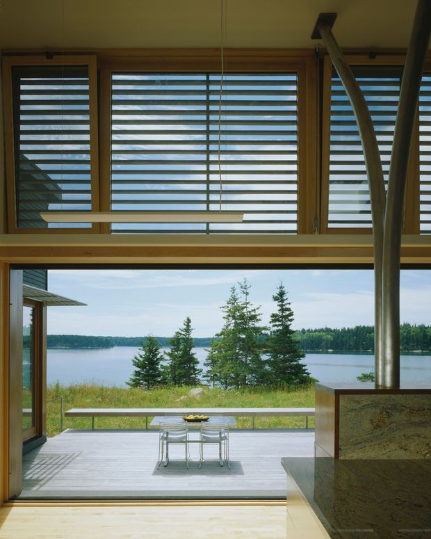 Interior view through large timber-framed glazing toward a serene lake and forested shoreline. Horizontal louvred or shuttered windows occupy the upper portion, while a wide opening leads to a timber deck with a small outdoor dining table and chairs. A sculptural metal fireplace flue and stone-clad hearth structure frame the right side. The space has a warm, natural, highly connected-to-landscape character.