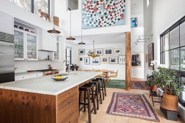 Bright, eclectic open-plan kitchen and dining area in a high-ceilinged interior. The space features a large white stone island with timber cabinetry and bar seating, white built-in kitchen cabinetry, pendant lights, a dining table, extensive framed artwork, a large abstract artwork above, exposed timber posts, tall industrial-style windows, indoor plants, and layered patterned rugs. A mezzanine or upper-level opening and clerestory glazing contribute to the airy vertical volume.