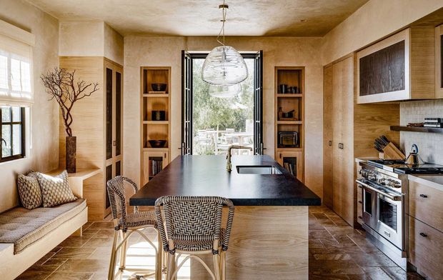 Warm, richly textured kitchen and dining space with a large dark island, integrated seating, timber cabinetry, open shelving, stainless-steel appliances, and a built-in window seat. Natural light enters through the windows and glazed rear doors, while textured plaster walls, timber joinery, and patterned furnishings create an inviting, refined atmosphere.