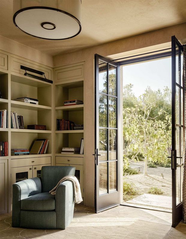 A warm, refined reading nook with built-in painted bookcases and lower cabinetry, filled with books and positioned beside open black-framed glazed French doors leading to a landscaped outdoor area. A rounded upholstered armchair, soft throw, neutral carpet, textured walls and a large circular ceiling light create a calm, inviting atmosphere.