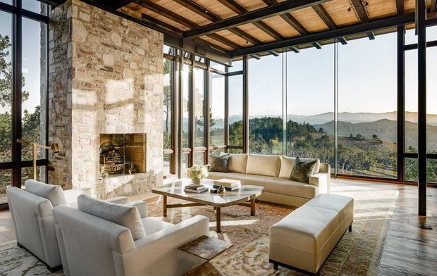 A spacious, light-filled living room with floor-to-ceiling glazing overlooking a wooded, mountainous landscape. The room features a large rough natural-stone fireplace, exposed timber ceiling beams and decking, dark slender structural frames, pale upholstered sofas and armchairs, a low timber-and-glass coffee table, and a large patterned rug. The interior has a refined contemporary lodge-like character with strong indoor-outdoor views.