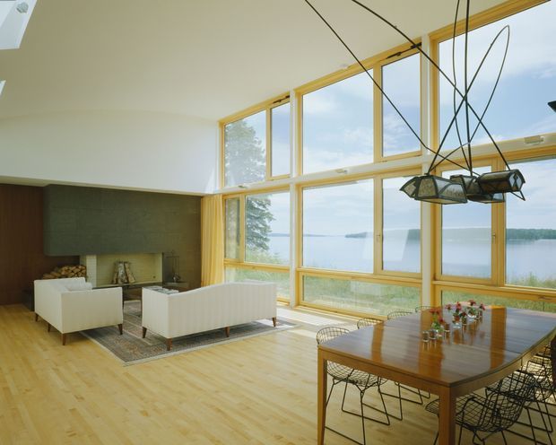 Bright open-plan living and dining room with expansive floor-to-ceiling timber-framed glazing overlooking a calm body of water. The room has a high sloping ceiling, pale timber flooring, white sofas, a dark feature wall with a fireplace, and a timber dining table with wire-frame chairs. A sculptural multi-arm pendant light hangs above the dining area.