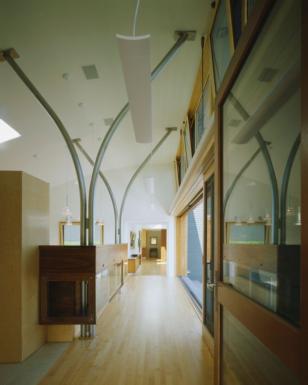 A bright, elongated modern interior corridor or entry passage with pale timber flooring, warm timber wall and door framing, extensive glazing, and built-in wood cabinetry. Sculptural curved metal ducts or structural supports rise from the cabinetry toward the ceiling, creating a distinctive architectural feature. Minimal white pendant lights and recessed ceiling fixtures illuminate the space, while a glass door and reflections are visible on the right.