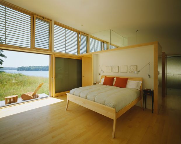 Bright bedroom with a low-profile timber bed, white bedding, and orange accent cushions. Full-height glazing and high horizontal louvred windows bring in abundant daylight and overlook a grassy waterfront landscape. Pale timber flooring, timber-framed glazing, and a partial timber room divider create a warm, airy, minimalist retreat.