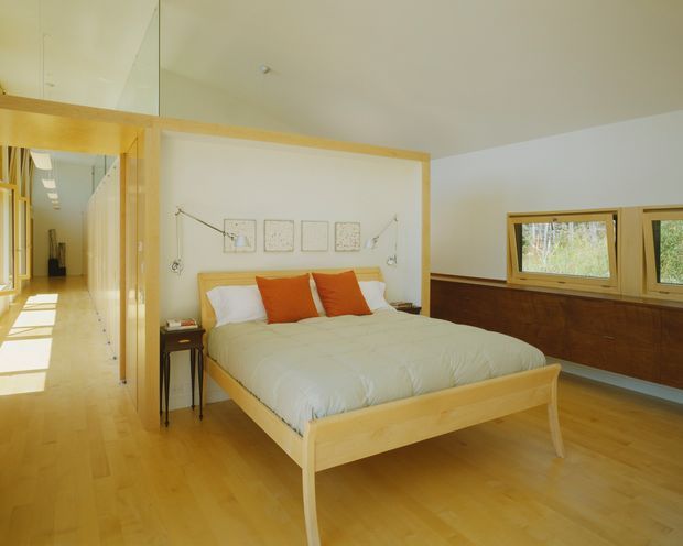 Bright, spacious bedroom with a light timber platform bed and matching timber flooring. The bed is dressed in white bedding with orange accent cushions, while a low dark timber built-in cabinet runs beneath a series of horizontal windows. A timber-framed opening leads to a long hallway, and simple wall-mounted reading lights and framed artwork add understated detail.