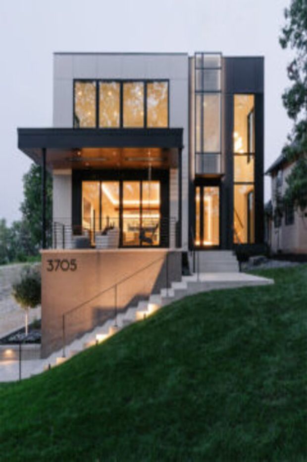 A dusk exterior view of a contemporary multi-storey residence on a steeply sloped site. The home features a rectilinear composition, flat rooflines, extensive floor-to-ceiling glazing, dark metal framing and cladding, pale rendered or panelled wall surfaces, and a projecting upper-level canopy. Illuminated interiors and stair lighting create a warm glow, while broad concrete steps and a low retaining wall lead up from the landscaped frontage.