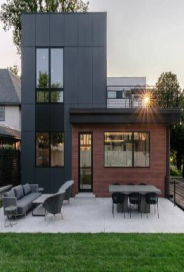 A contemporary multi-level residential extension viewed from the backyard. The home combines a tall dark charcoal cladded volume with narrow vertical glazing and a lower warm timber-clad pavilion. A rooftop terrace with glass or metal balustrading sits above the lower level, while a paved outdoor dining and lounge area occupies the foreground beside a lawn.