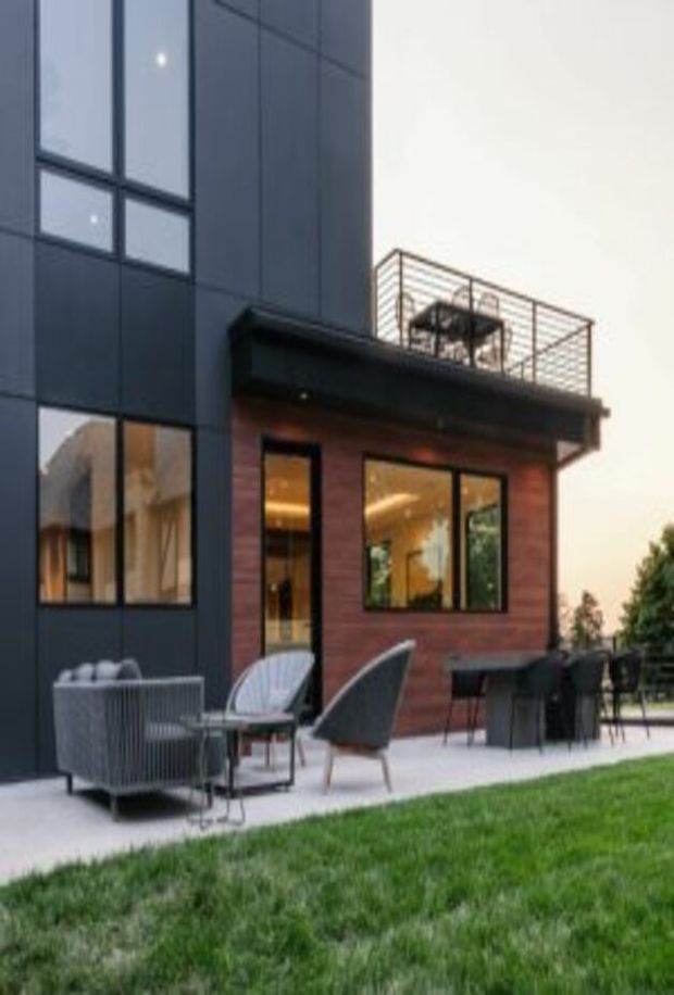 Contemporary multi-storey house exterior with a dark panelled upper facade, large black-framed windows, and a warm reddish brick lower-level extension. A covered ground-floor patio contains outdoor lounge and dining furniture, while a rooftop terrace with metal balustrading is visible above. The house opens onto a lawn and landscaped outdoor area in soft evening light.