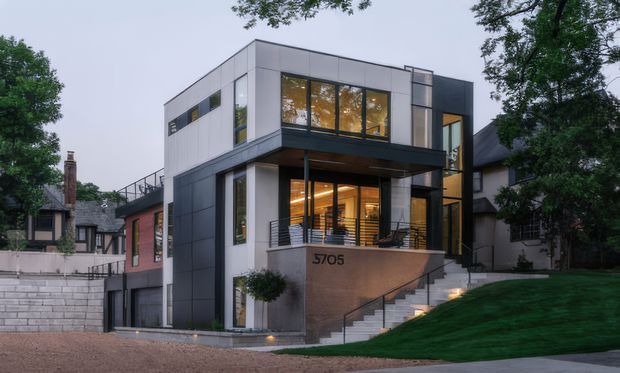 Exterior photograph of a contemporary multi-level urban residence set on a sloping landscaped site. The house has a strong rectilinear form, flat roof, white and dark grey panelled facade, expansive windows, a projecting upper volume, balcony-like terrace, exterior stairs, and a prominent concrete or stone entry wall marked 5705.