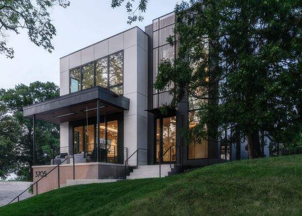 Exterior view of a contemporary multi-storey residence on a sloped, landscaped site. The building features crisp geometric volumes, large floor-to-ceiling windows, dark metal framing, pale panel cladding, and a broad projecting entry canopy. Steps and railings lead to a glazed entrance, with warm interior lighting visible through the windows and mature trees surrounding the property.