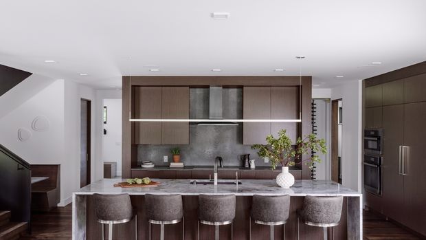 Contemporary kitchen viewed straight on, featuring a large marble-look island with four upholstered stools, dark timber cabinetry, a textured grey splashback, integrated stainless-steel appliances, and a linear pendant light. White walls and ceiling create a bright, refined interior, with a stair and adjacent circulation visible at the sides.
