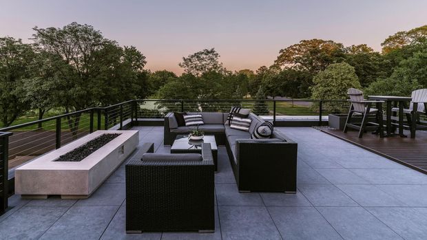 Expansive rooftop or elevated outdoor terrace furnished as an outdoor lounge and dining area. The space has large-format grey tiled paving, black metal balustrades, dark woven outdoor sofas and armchairs, a dining table with high-back chairs, and a long rectangular built-in fire feature. It overlooks a mature tree-filled landscape at dusk, creating a refined, tranquil entertaining setting.