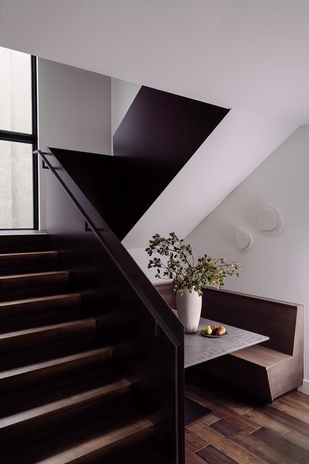 A refined modern stairway with dark timber treads, a dark sculptural balustrade, and a striking angular black geometric ceiling or wall feature. A tall window brings natural light into the space. Beside the stairs is a built-in dark timber bench with a grey stone-like table surface, decorative plant, and subtle white wall reliefs. The interior has a dramatic, sophisticated palette of dark wood, black, white, and grey.