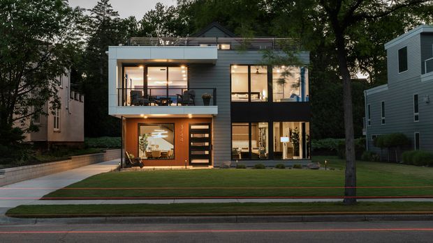 Evening exterior view of a modern multi-storey urban residence with a compact flat-roofed form, horizontal grey cladding, extensive floor-to-ceiling glazing, balconies, and a warm illuminated interior. The house sits behind a landscaped lawn with a paved driveway and mature trees.