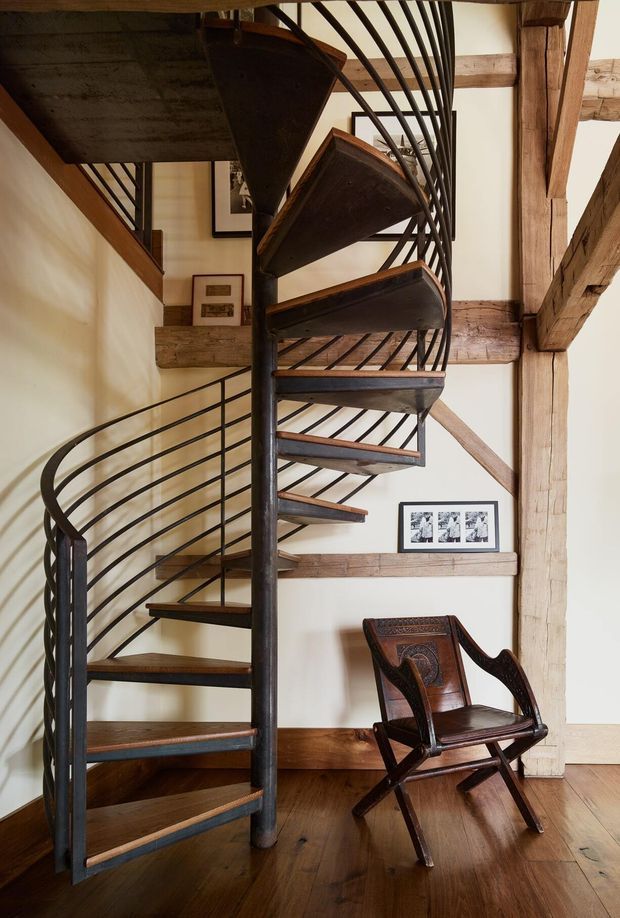 Interior view of a rustic timber-framed stairwell featuring a sculptural dark metal spiral staircase with timber treads and curved balustrade. Exposed heavy timber beams, warm wood flooring, cream plaster walls, framed artwork, and a carved wooden chair create a heritage-inspired, industrial-rustic atmosphere.