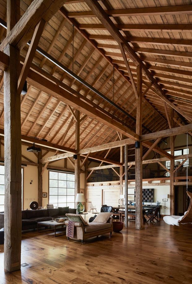 A spacious open-plan living area inside a barn-like timber structure, featuring dramatic exposed post-and-beam framing, a high pitched timber roof, broad polished wood flooring, large industrial-style grid windows, a dark sofa, leather lounge chairs, dining furniture, and a mezzanine-level railing. The interior combines rustic structural character with comfortable contemporary furnishings.