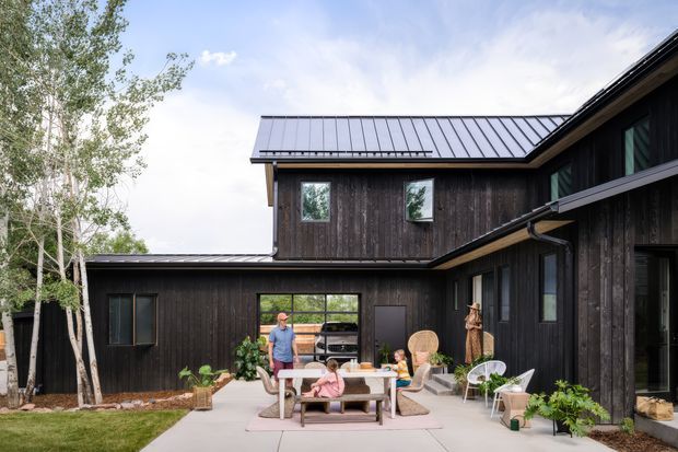 Exterior view of a dark-clad multi-wing home arranged around a paved courtyard patio. The house features vertical timber cladding, large glazed openings, and a standing-seam metal gable roof, with outdoor dining furniture and planting in the foreground.