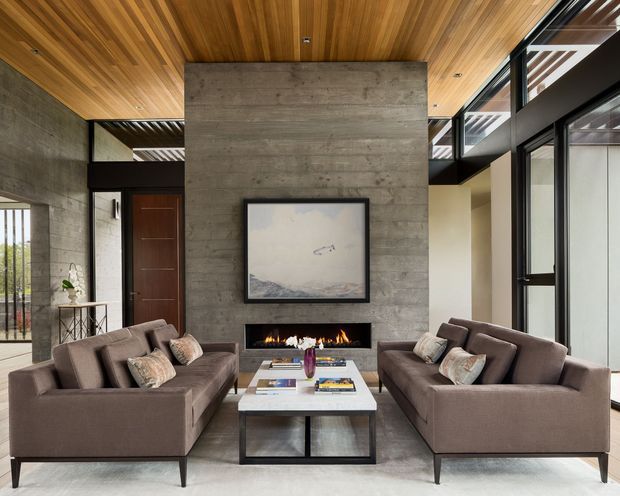 A refined contemporary living room with two facing taupe sofas arranged around a low rectangular coffee table. The room features a dramatic exposed concrete feature wall with a linear fireplace and artwork, a warm timber slatted ceiling, dark-framed full-height glazing, and an adjacent entry door. The balanced symmetrical layout and layered natural materials create a sophisticated, warm atmosphere.