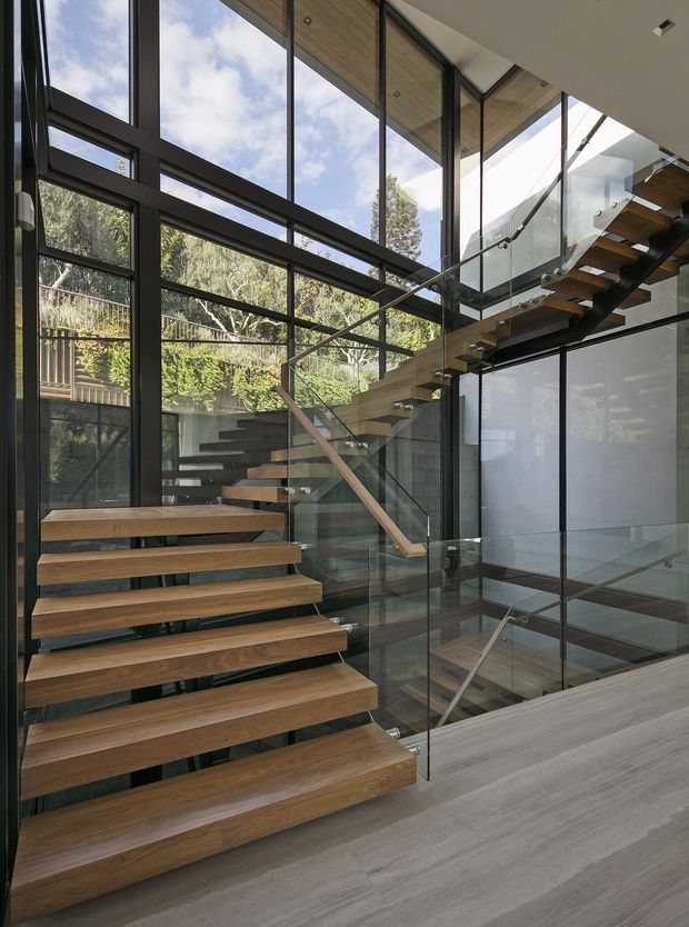 A contemporary interior stairway with floating timber treads, slim dark metal supports, and clear glass balustrades. Floor-to-ceiling glazing surrounds the stair, bringing views of leafy vegetation inside, while timber ceiling lining and dark-framed windows reinforce the modern architectural character. The stair appears to connect multiple levels around a void, with strong natural light and open visual flow.