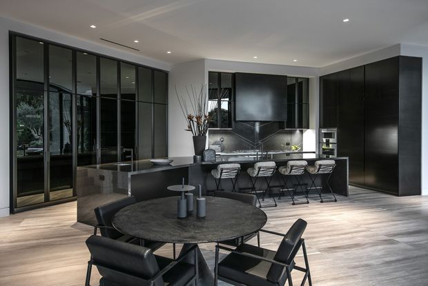 A sophisticated open-plan kitchen and dining area with dark, full-height cabinetry, a large central island with bar seating, and a round dining table with black chairs. Black-framed glazed partitions and doors create visual separation while maintaining openness. Light timber flooring, pale walls, recessed ceiling lighting, and reflective dark surfaces produce a refined, contemporary monochrome interior.