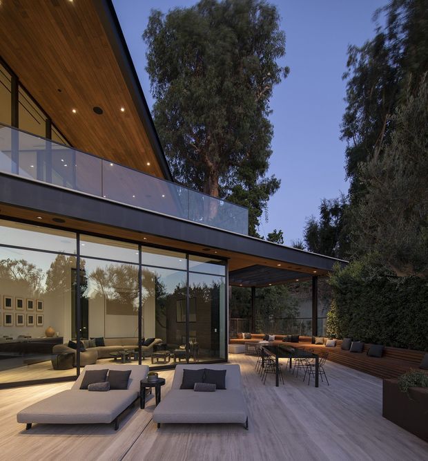 A refined modern courtyard terrace viewed at dusk, with extensive floor-to-ceiling glazing opening from a living area onto a timber deck. The outdoor space includes low-profile lounge seating, a dining table, built-in bench seating, planting, and a transparent glass balustrade. A dramatic timber-lined soffit and broad cantilevered roof create a strong indoor–outdoor connection, with mature trees surrounding the property.