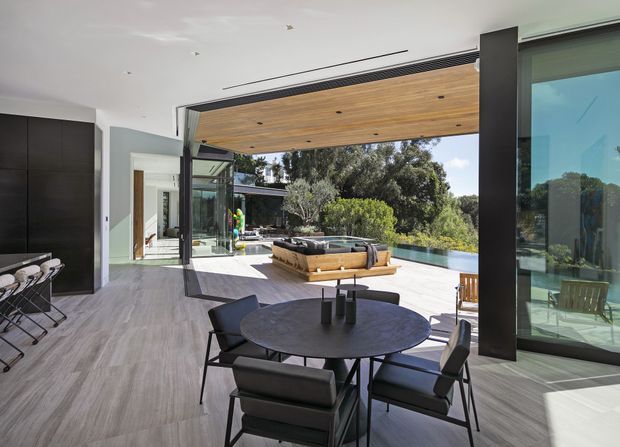 A contemporary open-plan indoor-outdoor living and dining area with a round dark dining table and upholstered chairs in the foreground. Large retractable glazed openings connect the room to a spacious terrace with outdoor seating and a swimming pool beyond. The space features a warm timber-lined ceiling, pale wood-look flooring, dark built-in cabinetry, slim black structural columns, and extensive floor-to-ceiling glazing, creating a bright, seamless connection to the landscaped setting.