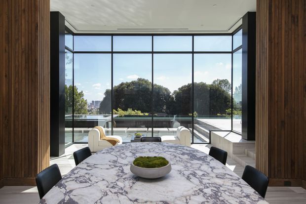 A sophisticated dining area with a large rectangular marble dining table and dark upholstered chairs, framed by warm vertical timber wall panelling. Floor-to-ceiling black-framed glazing opens to a bright terrace or poolside setting, with sculptural cream lounge chairs and a contemporary linear ceiling detail. The space has a refined, high-contrast modern character with strong indoor-outdoor connection.
