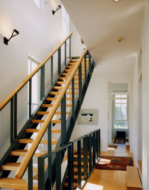 Bright multi-level stairway and landing in a tall, narrow interior. The staircase has open timber treads, warm timber handrails, and dark painted vertical metal balustrades. White walls, tall windows, recessed ceiling lights, timber flooring, and patterned rugs create a clean, airy transitional space.