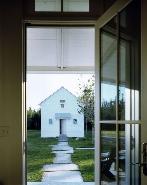 View from inside through an open glazed door toward a simple white weatherboard outbuilding with a steep gabled roof. A concrete stepping-stone path crosses a neatly maintained lawn, while the doorway and surrounding interior are dark and framed by timber elements. The scene has a restrained, rural architectural character with strong indoor–outdoor connection.