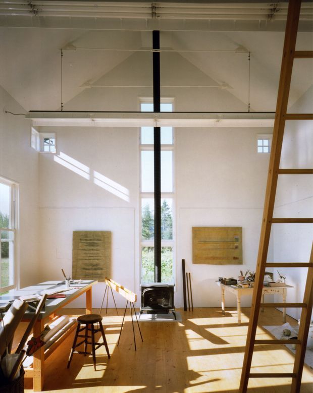 A bright, double-height artist's studio or creative workspace with white walls, exposed structural roof framing, a central black flue and freestanding wood-burning stove, tall multi-pane windows, timber floors, worktables, canvases, easels, stools, and a prominent timber ladder leading to an upper level or loft. Strong sunlight creates dramatic diagonal shadows across the floor and walls.