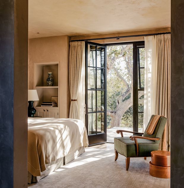 A warm, refined bedroom viewed through a wide doorway, featuring a neatly made bed, built-in recessed shelving and cabinetry, a vintage-style green upholstered armchair with timber arms, a small wooden side table, full-height cream curtains, and black-framed glazed French doors opening to leafy trees outside. Soft sunlight creates dappled shadows across the bed and carpet.