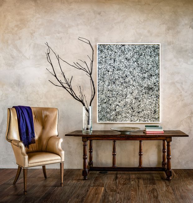 A refined traditional-style interior vignette featuring a tan tufted leather wingback chair with a draped deep-purple throw, positioned beside a dark timber console table. The console displays a glass vase with bare branches, a shallow decorative bowl, and stacked books beneath a large framed textured artwork. The room has a warm mottled plaster wall and dark richly grained timber flooring.