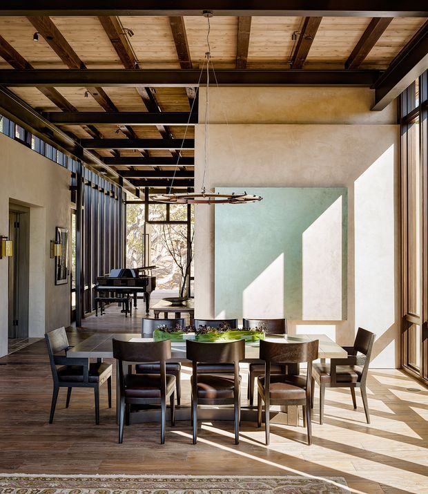 A spacious, sunlit dining room with a long dark wood dining table and upholstered dining chairs, opening toward a living area with a grand piano. The room features expansive floor-to-ceiling glazing, textured plaster walls, exposed dark timber beams, a pale timber ceiling, and a sculptural linear pendant light. Strong geometric sunlight and shadows fall across the pale wall and timber floor, creating a warm, refined atmosphere.