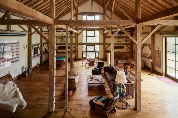 Open-plan rustic interior in a converted barn-like structure, featuring exposed timber post-and-beam framing, exposed rafters, pitched roof, wide timber flooring, built-in shelving, and a combined living and dining area. Large multi-pane windows and glazed doors provide abundant natural light. Leather and upholstered seating, a central coffee table, and warm timber finishes create a relaxed, inviting atmosphere.