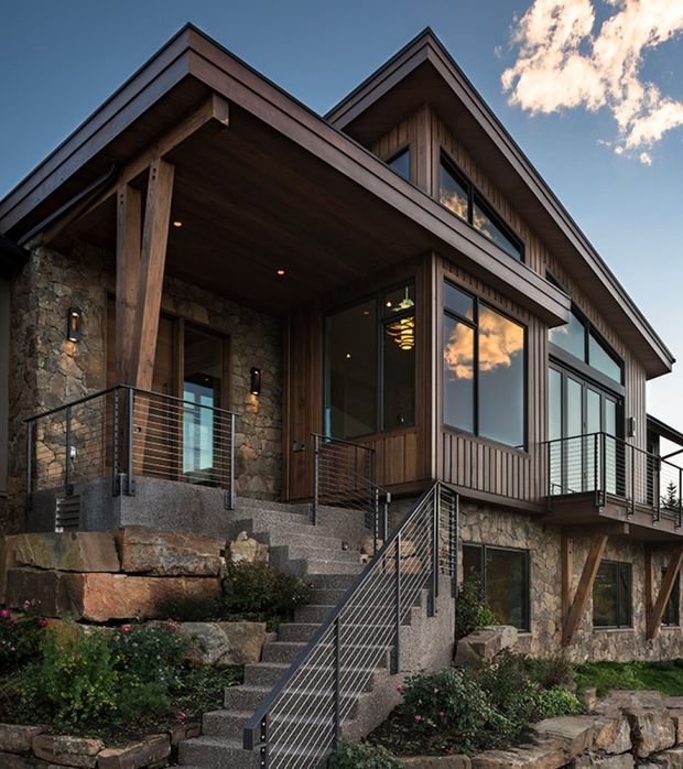 Exterior view of a multi-level mountain-style home with steep gabled rooflines, extensive timber cladding and soffits, natural stone walls, large dark-framed windows, and projecting balconies. Broad exterior stairs and metal cable railings connect the landscaped, rocky site to the covered entry porch.