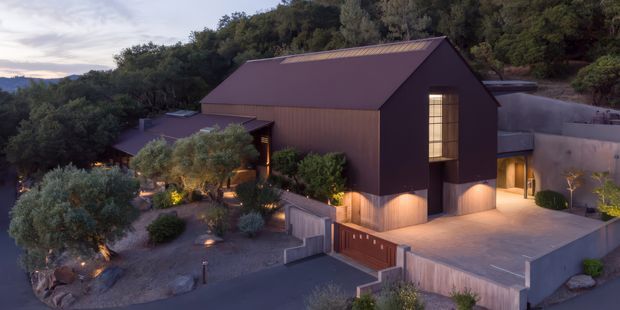 Twilight exterior view of a contemporary, multi-level residence set into a wooded, sloping site. The home features a large dark gabled volume with vertical cladding, warm illuminated glazing, adjoining flat-roofed forms, concrete retaining walls, and landscaped terraces with mature olive trees and integrated garden lighting.